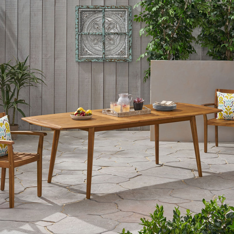 Outdoor Noble House Placidity expandable dining table with slatted tabletop and tapered legs, shown in a spacious outdoor setting.