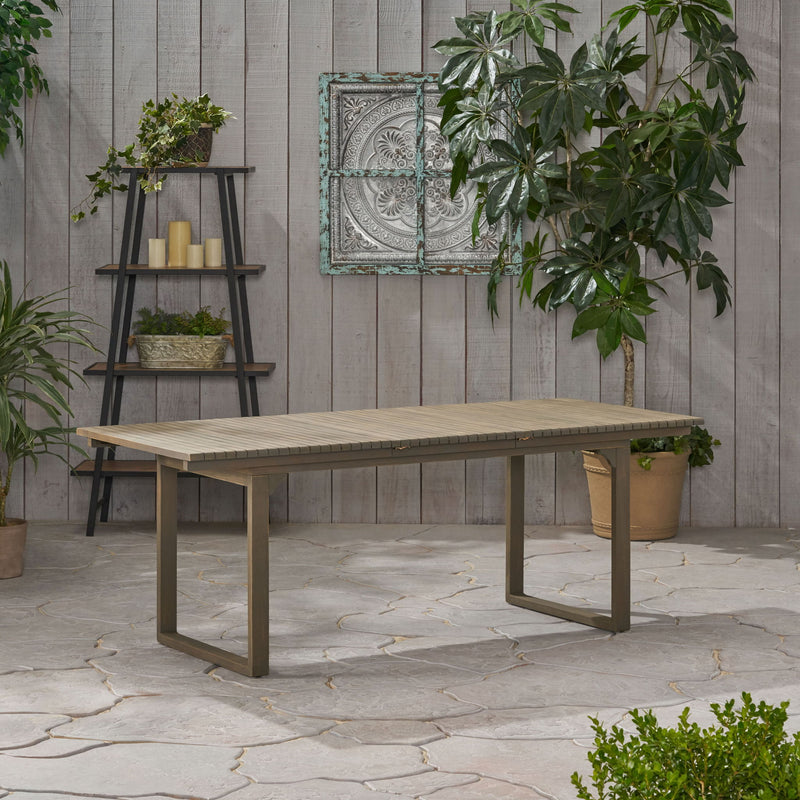 Outdoor expandable acacia wood dining table with U-shaped legs by Noble House, showing the table extended with a natural wood finish.