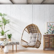 Light brown wicker hanging chair with beige cushion and 8-foot chain, ideal for outdoor or indoor relaxation.