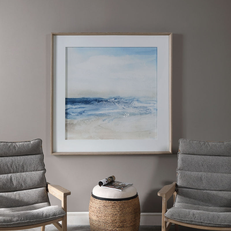 Framed print titled "Surf And Sand" by Uttermost featuring a serene beach scene with blue tones and a stylish frame.